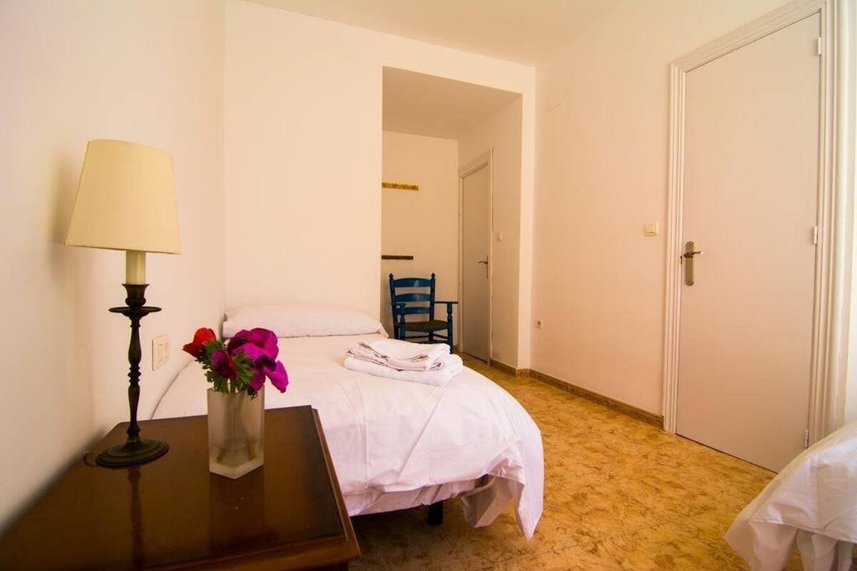A hotel room with a bed, a bedside table with a lamp and flowers, a chair, and a door.