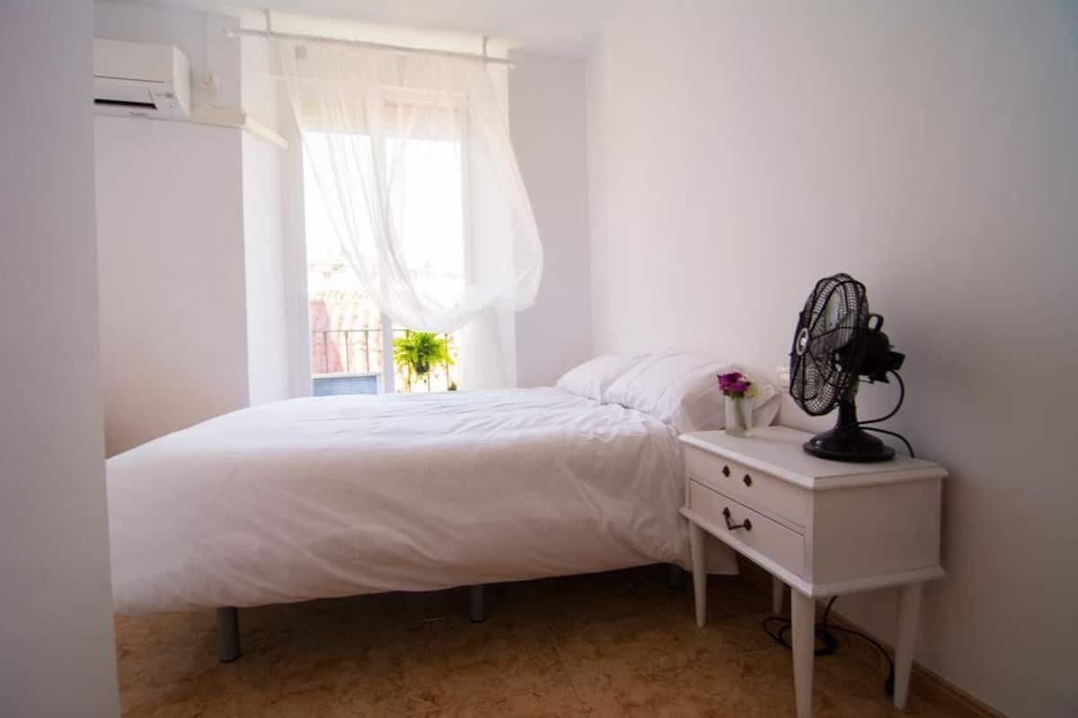 A bedroom with a bed, a bedside table with a fan, and a window with sheer curtains.