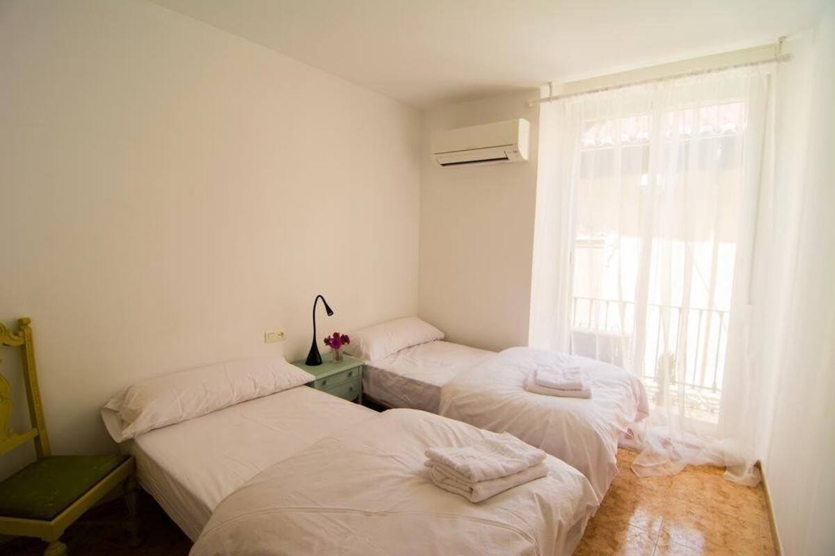 A room with two beds, a small table with a lamp, and a window with sheer curtains.