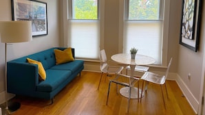 A room with a blue sofa, a small table with chairs, a floor lamp, and two windows with blinds. - Dupont 5-Star, Walk Score 94, Metro, 2BR (Washington)