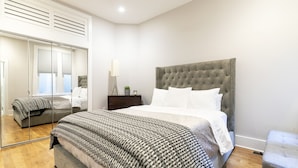 A modern bedroom with a large bed, a grey upholstered headboard, a bedside table with a lamp, and a mirror cabinet. - Dupont 5-Star, Walk Score 94, Metro, 2BR (Washington)