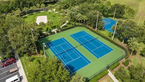 Vue aérienne d’un complexe de courts de tennis, entouré d’une végétation luxuriante et d’immeubles d’habitation.