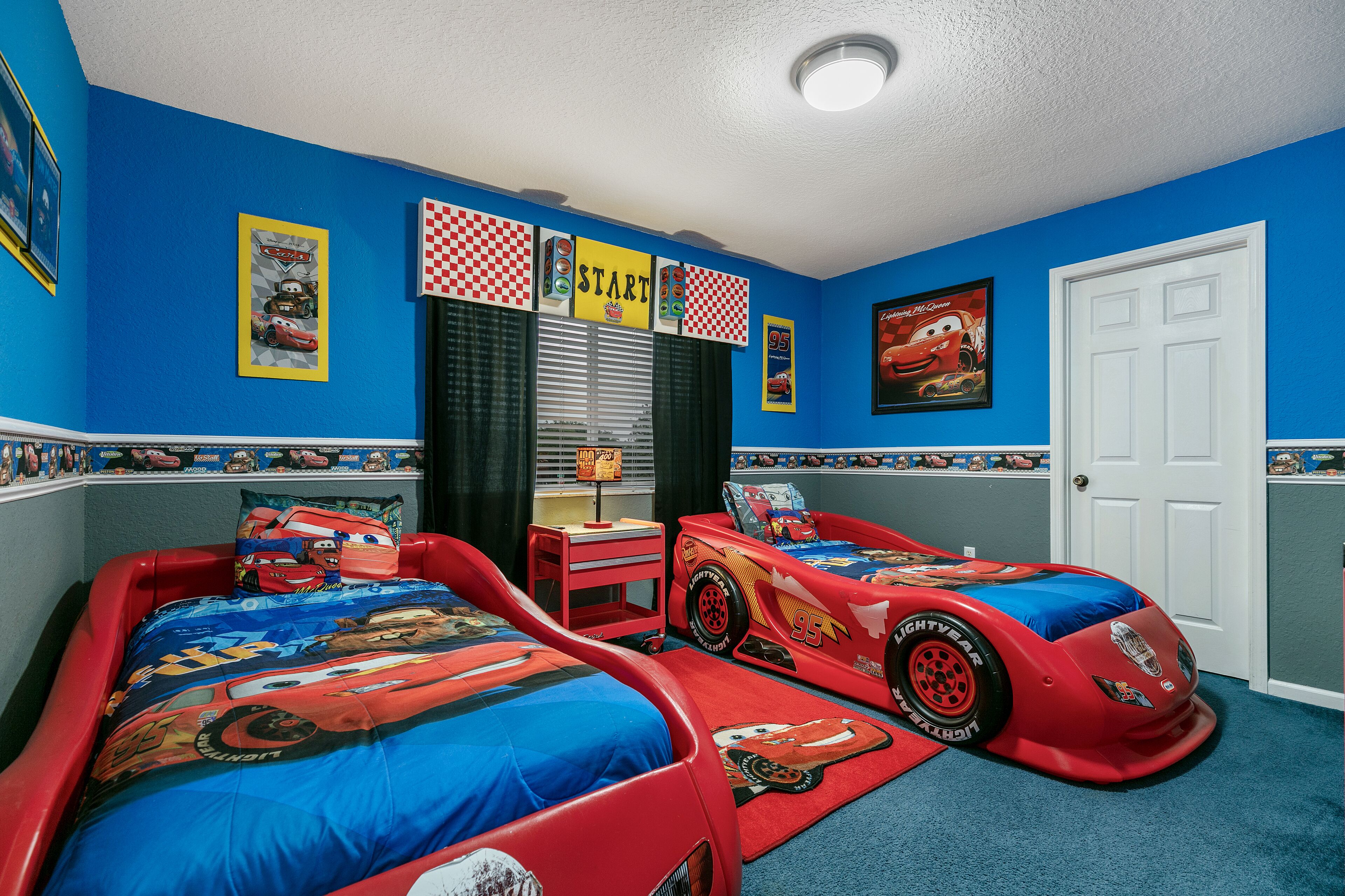 Une chambre avec deux lits, chacun agrémenté d’une couvre-lit sur le thème de l’automobile, un bureau et une fenêtre avec des stores.