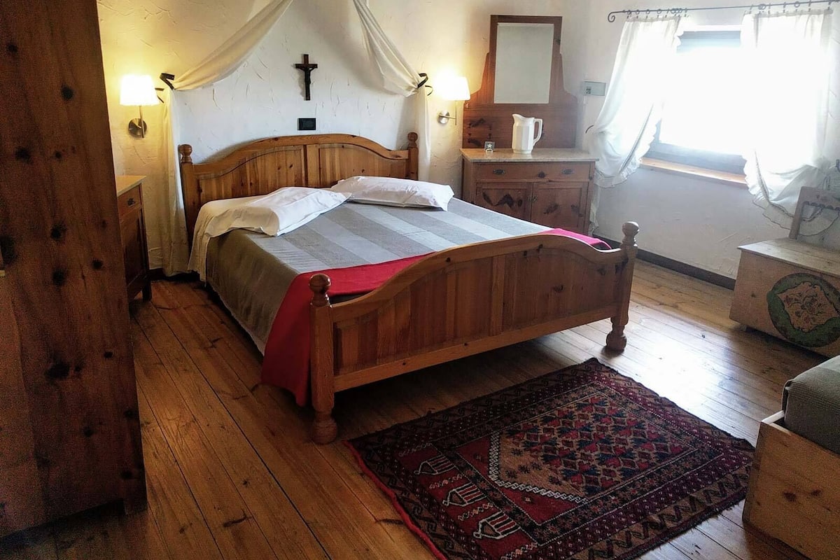 A wooden bed with a red blanket, a wooden nightstand, a mirror, and a wall-mounted cross.