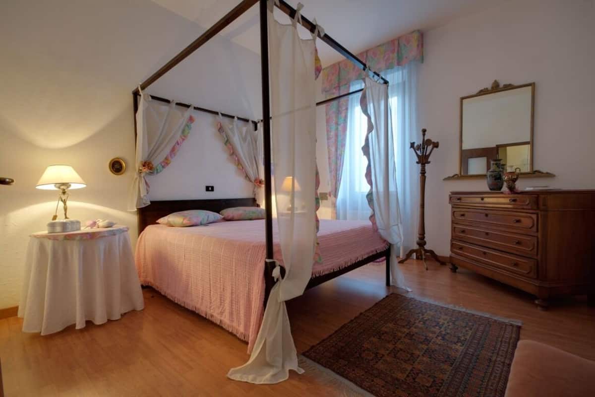 A bedroom with a four-poster bed, a bedside table with a lamp, a dresser, and a mirror on the wall.