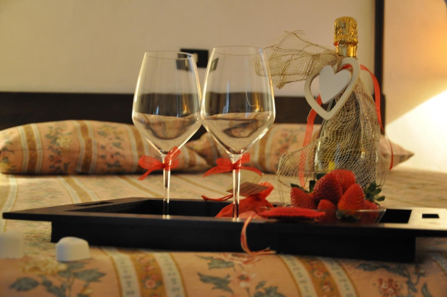 A tray with two wine glasses, a bottle of champagne, and a heart-shaped decoration on a bed with floral-patterned bedding.