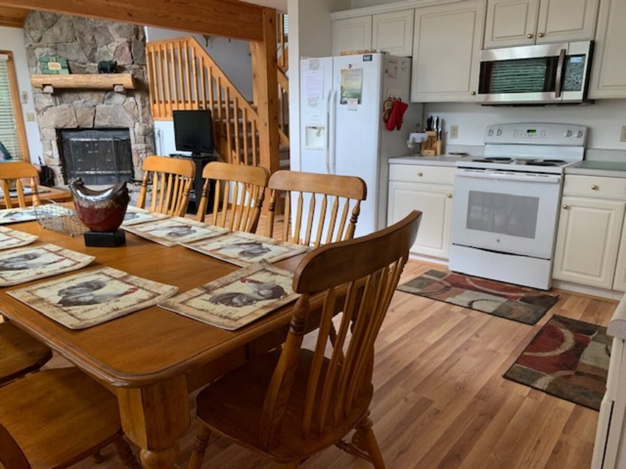 Un comedor con una mesa de madera preparada para cuatro personas, una chimenea de piedra y una cocina con electrodomésticos blancos.