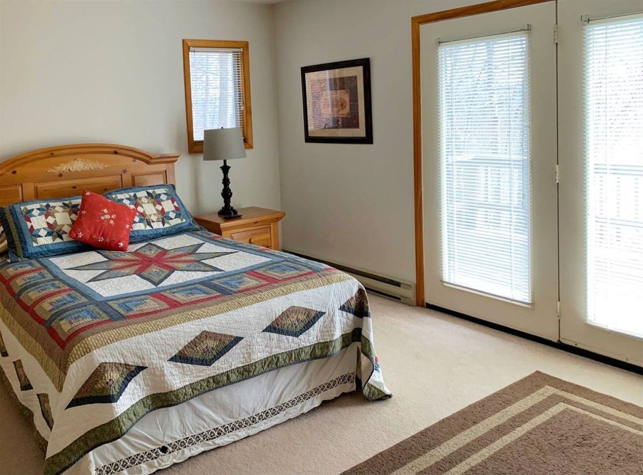 A bedroom with a bed, a nightstand, a lamp, a framed picture, and a window with blinds.