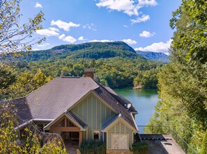 A house with a sloped roof, a stone chimney, and a view of a lake surrounded by forested hills. - Over the Edge - Lakefront, Hot tub, Sleeps 10, Two kayaks (Lake Lure)
