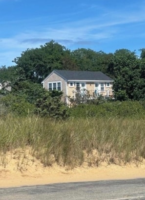 A two-story house with a gabled roof, surrounded by dense greenery and a sandy beach.