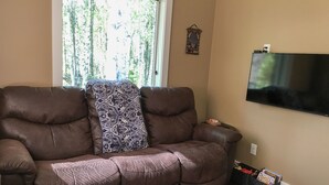 A brown leather sofa with a patterned throw pillow, a flat-screen TV mounted on the wall, and a small side table with remote controls. - Talkeetna's Mount Dall Cottage - WiFi&laundry - easy access from Parks Highway! (Talkeetna)