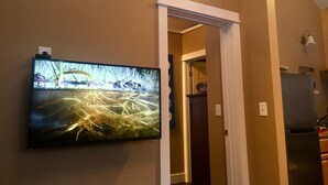 A room with a television mounted on the wall displaying a nature scene. There is a doorway leading to another room, and a refrigerator is visible in the background. - Talkeetna's Mount Dall Cottage - WiFi&laundry - easy access from Parks Highway! (Talkeetna)
