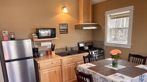 A compact kitchen with a stainless steel refrigerator, a microwave, a stove, and wooden cabinets. There's a dining table with a tablecloth and chairs. - Talkeetna's Mount Dall Cottage - WiFi&laundry - easy access from Parks Highway! (Talkeetna)
