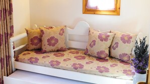 Una habitación acogedora con un sofá cama blanco, cojines con estampado floral, techo de madera, aire acondicionado montado en la pared, ventana con marco de madera y una pequeña planta en maceta.