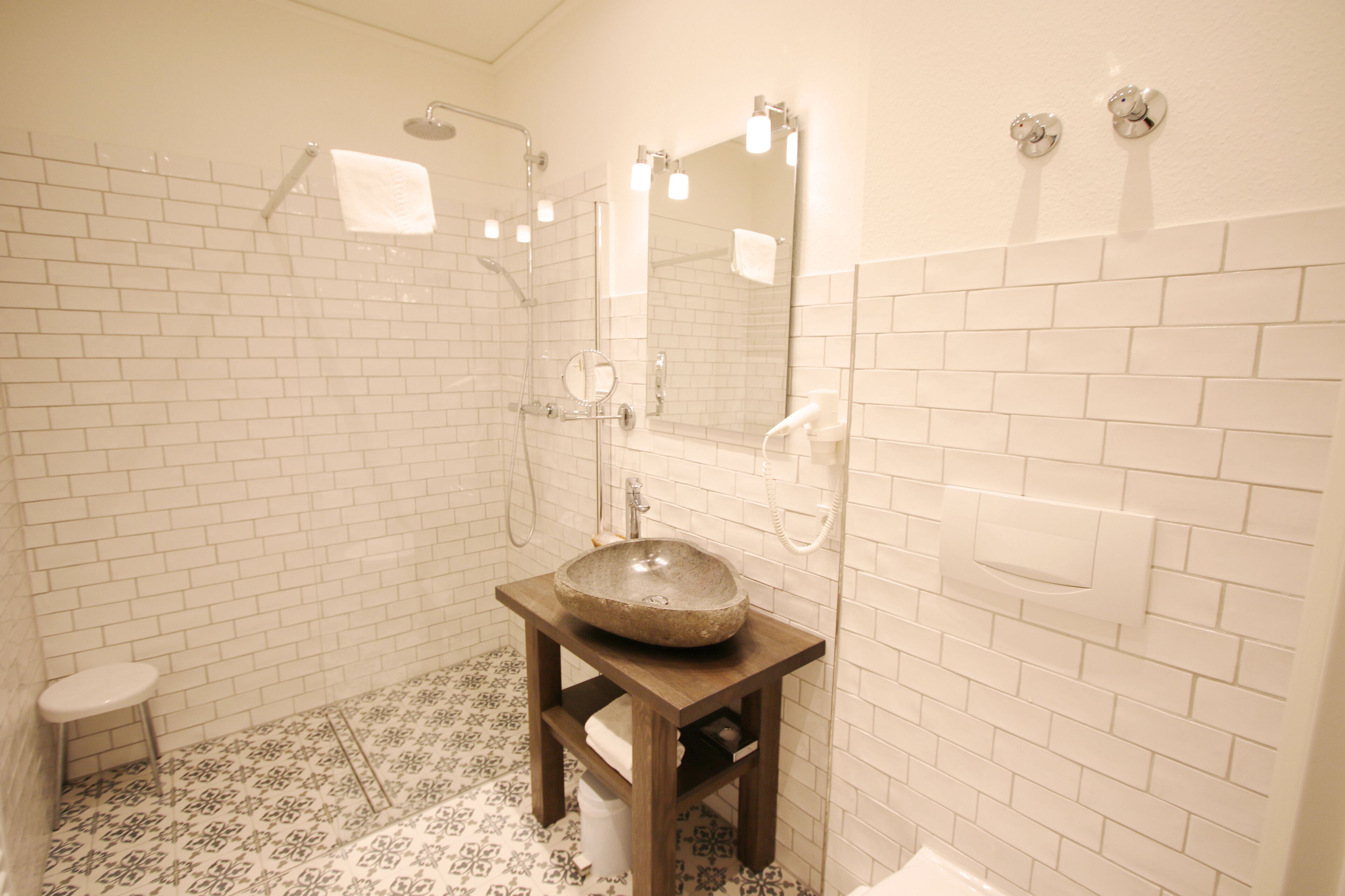 Een badkamer met witte betegelde wanden, een houten wastafelmeubel met een stenen wastafel, een handdouche en een klein krukje.