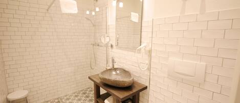 Un baño con paredes de azulejos blancos, un mueble de tocador de madera con lavabo de piedra, un cabezal de ducha manual y un taburete pequeño.