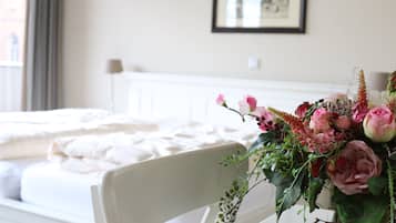 A hotel room with a neatly made bed, a white chair, and a floral arrangement on the side table.