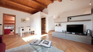 A spacious living room with a wooden ceiling, a large flat-screen TV, a glass coffee table with a book, and a modern kitchen in the background.