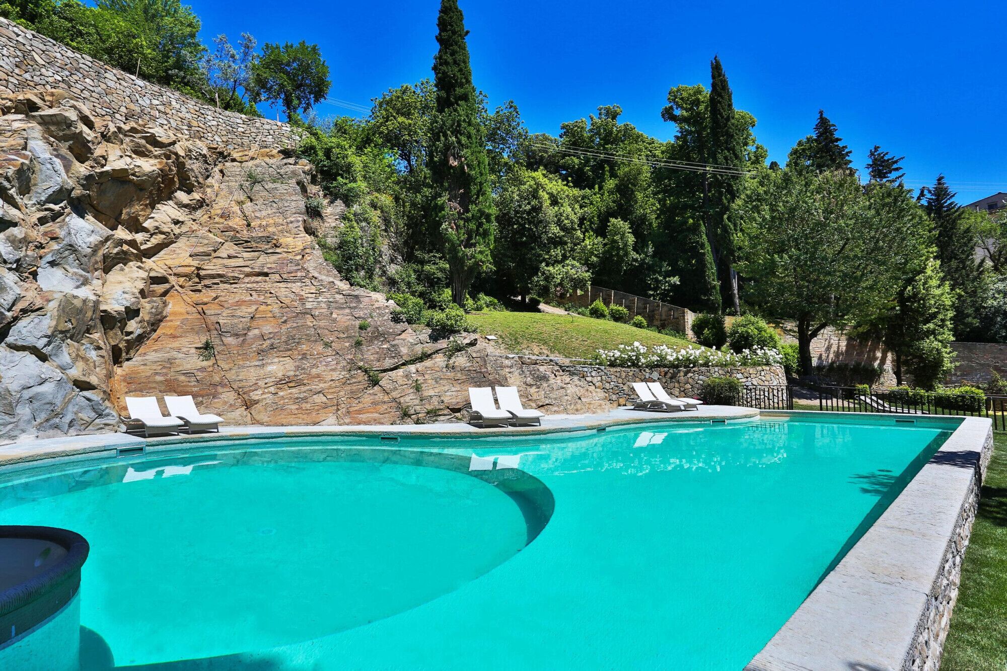Una piscina con sillones de descanso, un muro de piedra y árboles al fondo.