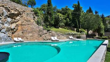 Una piscina con sillones de descanso, un muro de piedra y árboles al fondo.