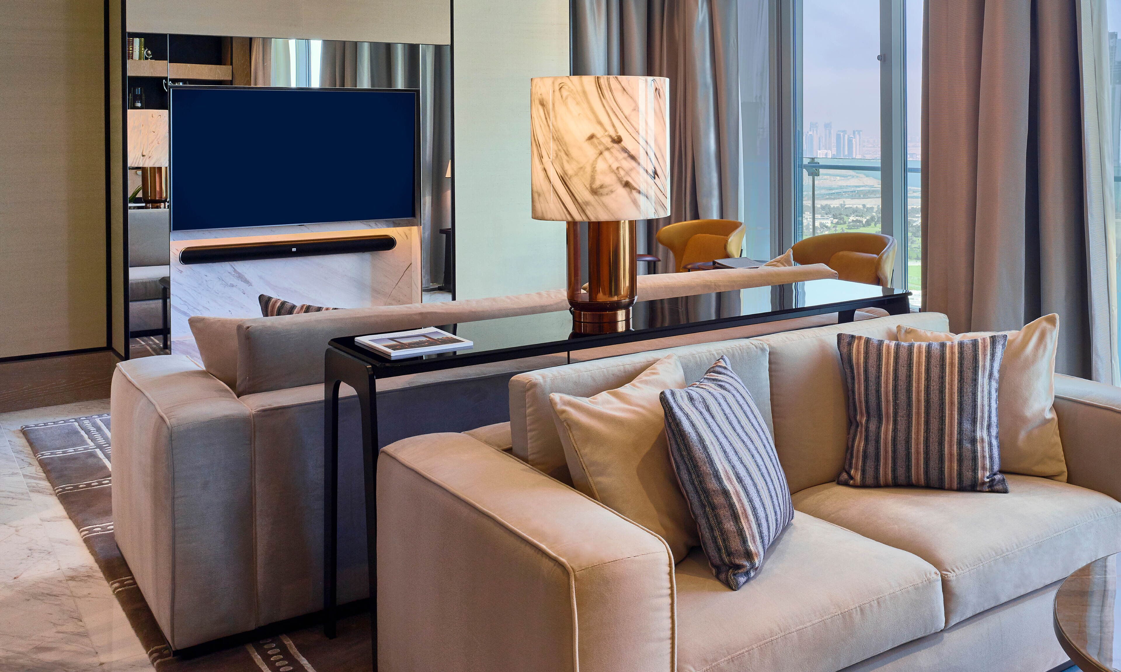 A modern hotel room with a large flat-screen TV, a sofa, a coffee table, and a lamp.