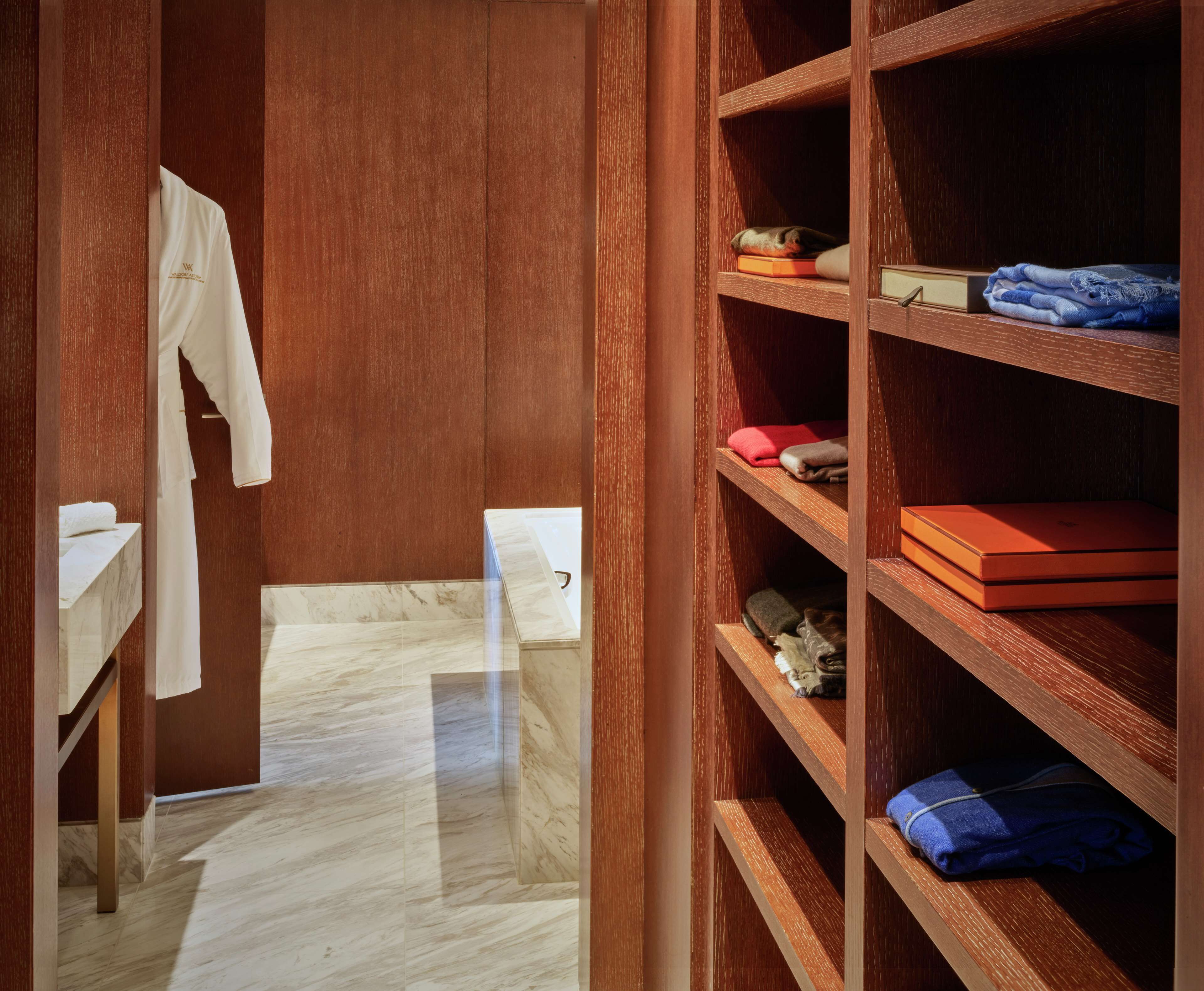 Un baño con piso de mármol, una bata blanca colgada y repisas de madera con toallas dobladas y una caja roja.