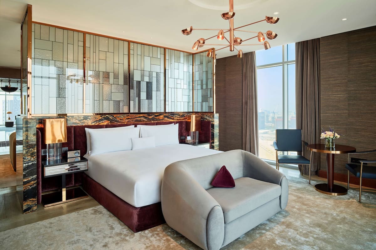 A modern hotel room with a large bed, a seating area, a desk, and a view of the city.