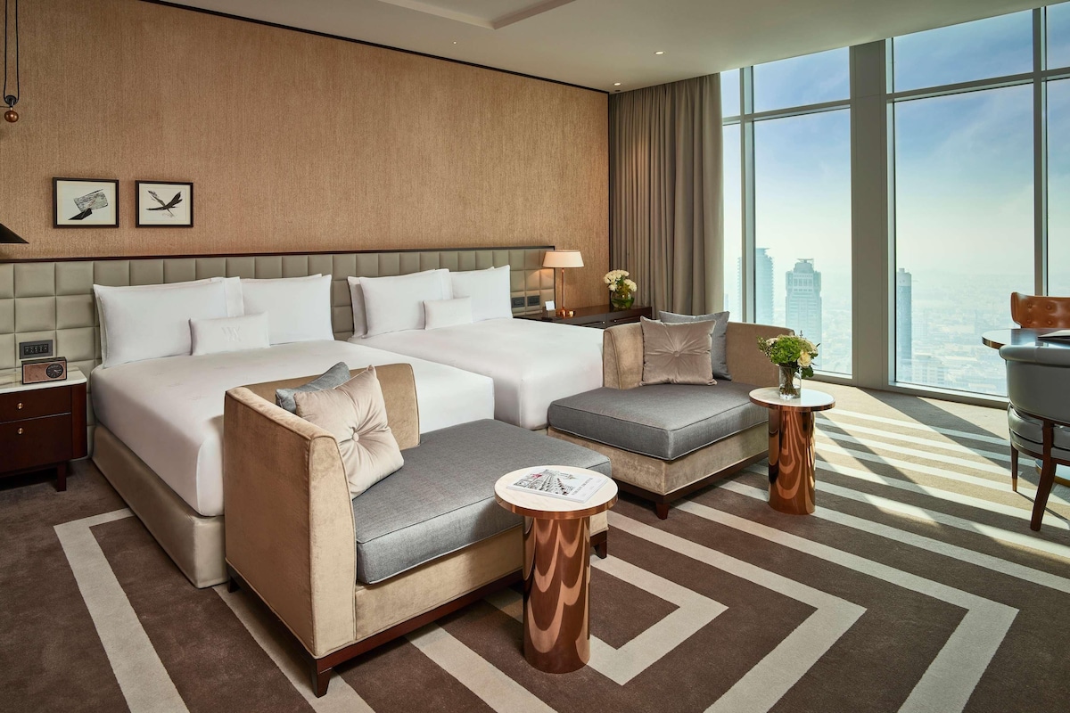 Una habitación de hotel moderna con una cama grande, un sofá, una mesita y vistas a la ciudad a través de amplios ventanales.