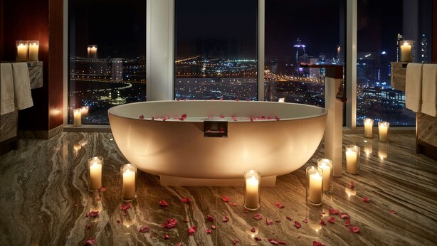 Un baño moderno con una bañera independiente, múltiples velas y pétalos de rosa sobre un suelo de mármol, con vistas a un paisaje urbano nocturno.