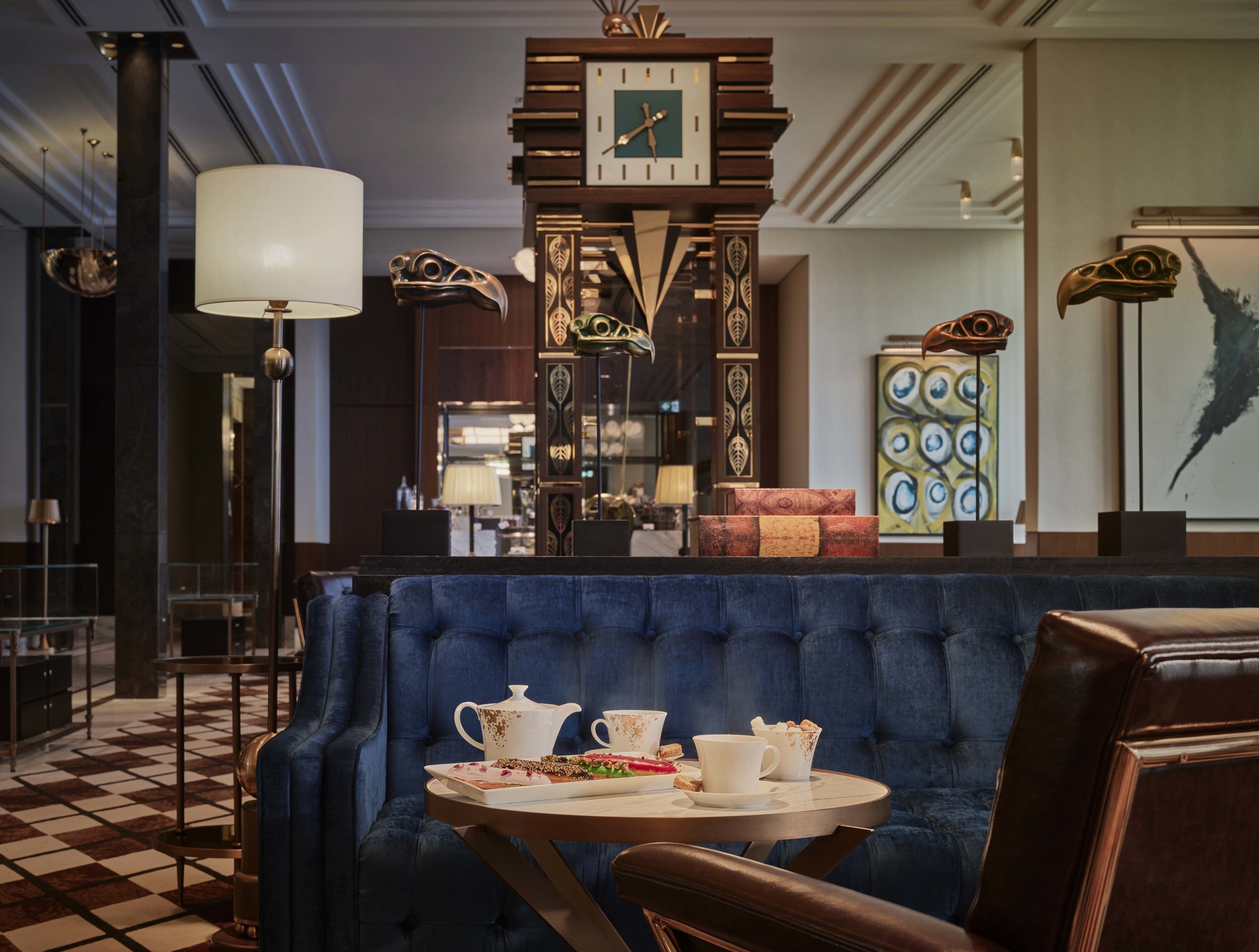 A lobby with a blue upholstered bench, a round table with a teapot, cups, and a plate of food, a large wall clock, and decorative items including a sculpture and framed artwork.