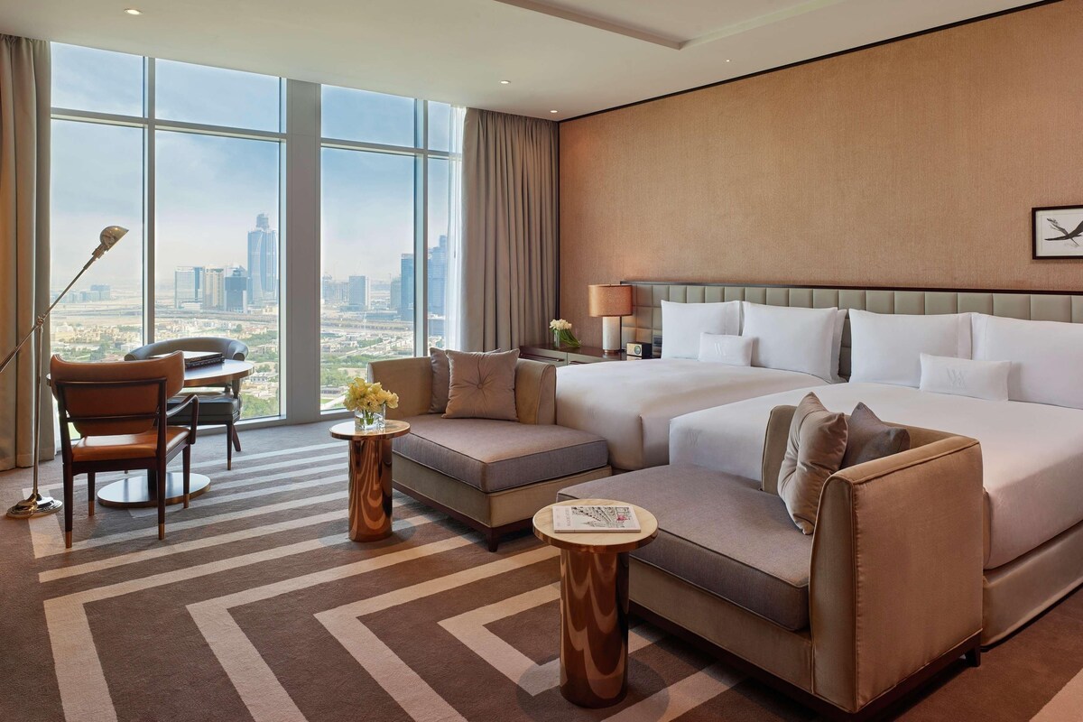 A hotel room with a large bed, a sofa, a chair, and a desk. There is a view of the city through large windows.