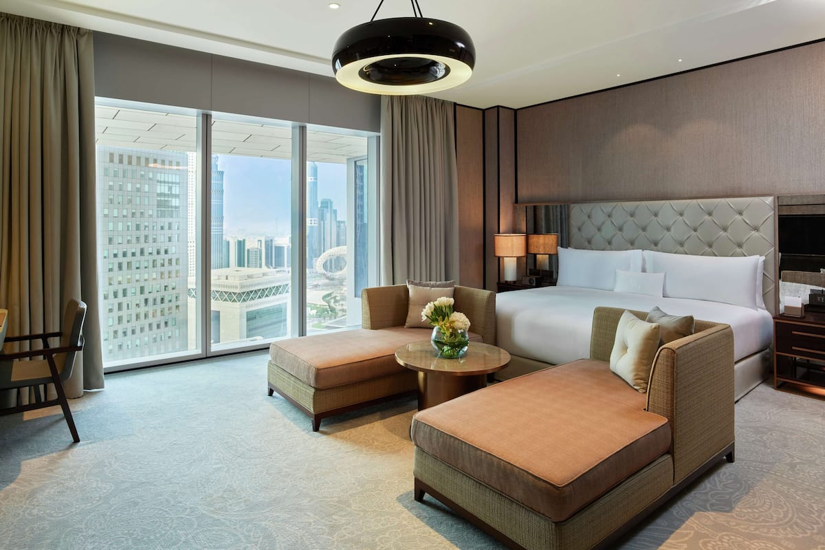 A modern hotel room with a large bed, two sofas, a dining table, and a view of the cityscape through floor-to-ceiling windows.