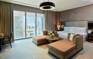 A modern hotel room with a large bed, two sofas, a dining table, and a view of the cityscape through floor-to-ceiling windows.