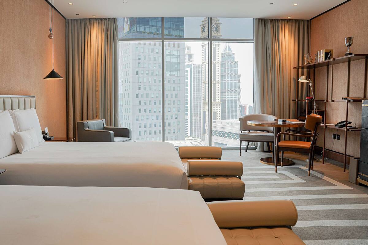 A hotel room with two beds, a desk, a chair, a sofa, and a view of city buildings.