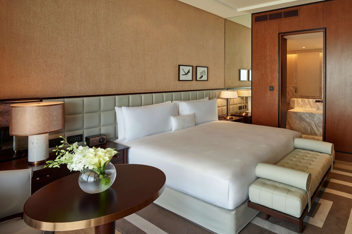 A hotel room with a large bed, a bedside table with a lamp, a small table with a vase of flowers, and a seating area with a sofa and armchairs.