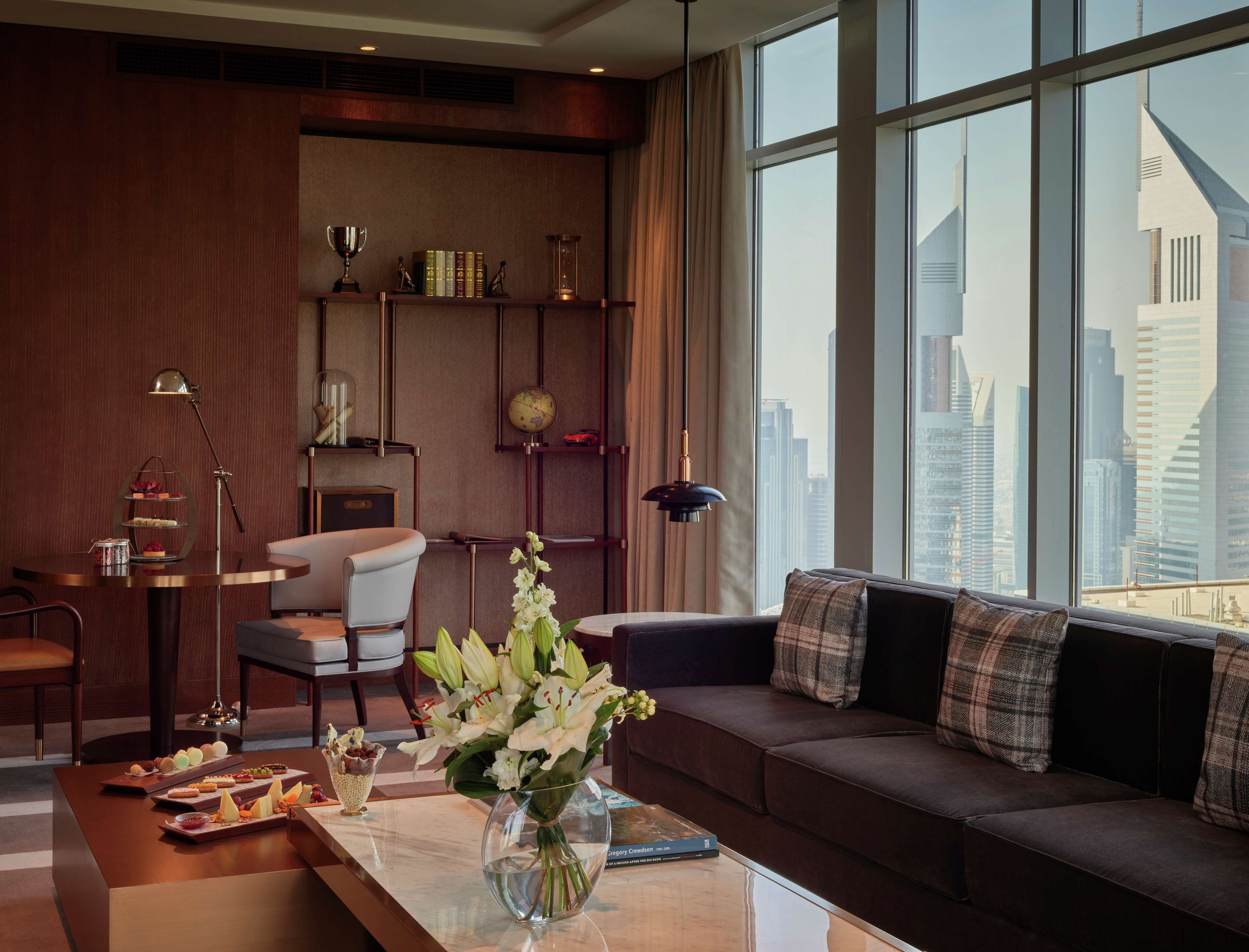 A modern living room with a sofa, a coffee table with desserts, a bookshelf, and a city view through large windows.