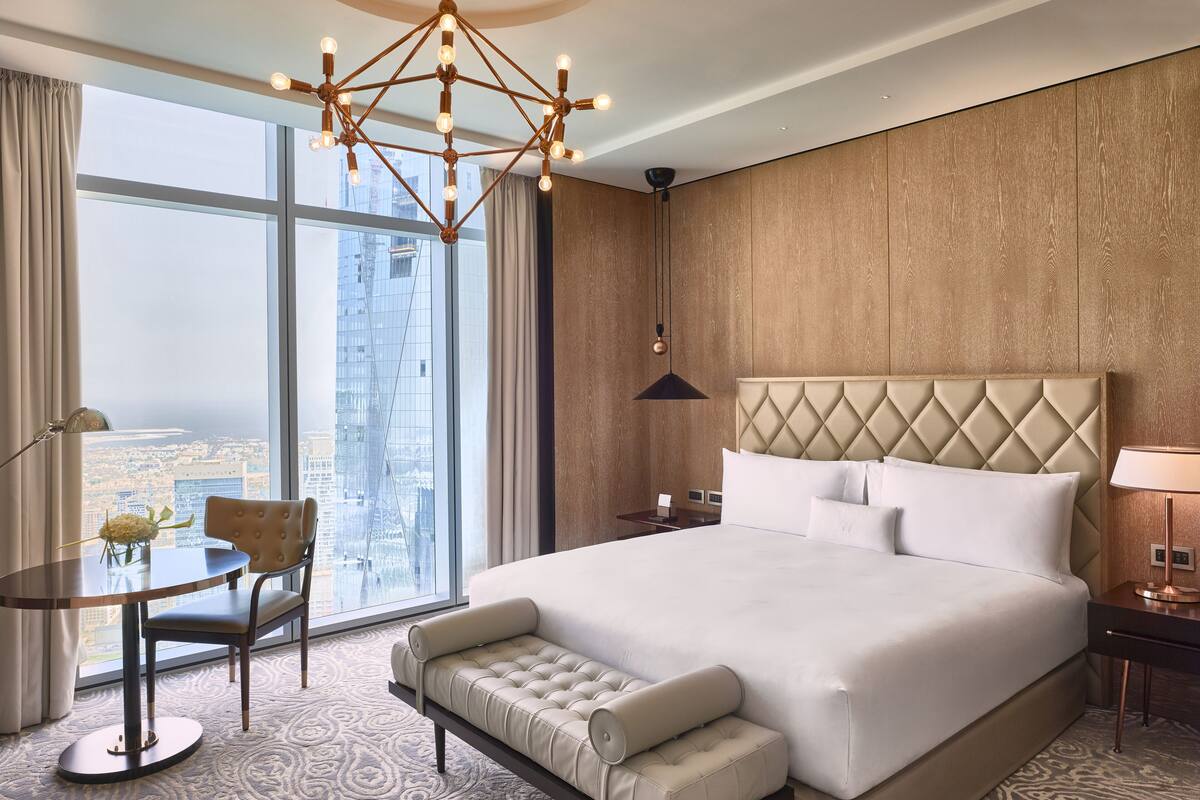 A modern hotel room with a large bed, a seating area, a desk, and a view of a cityscape through large windows.