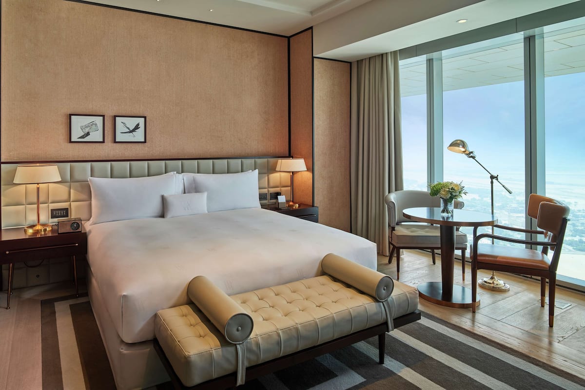 A hotel room with a large bed, bedside lamps, a small table with a chair, and a view of the city through large windows.