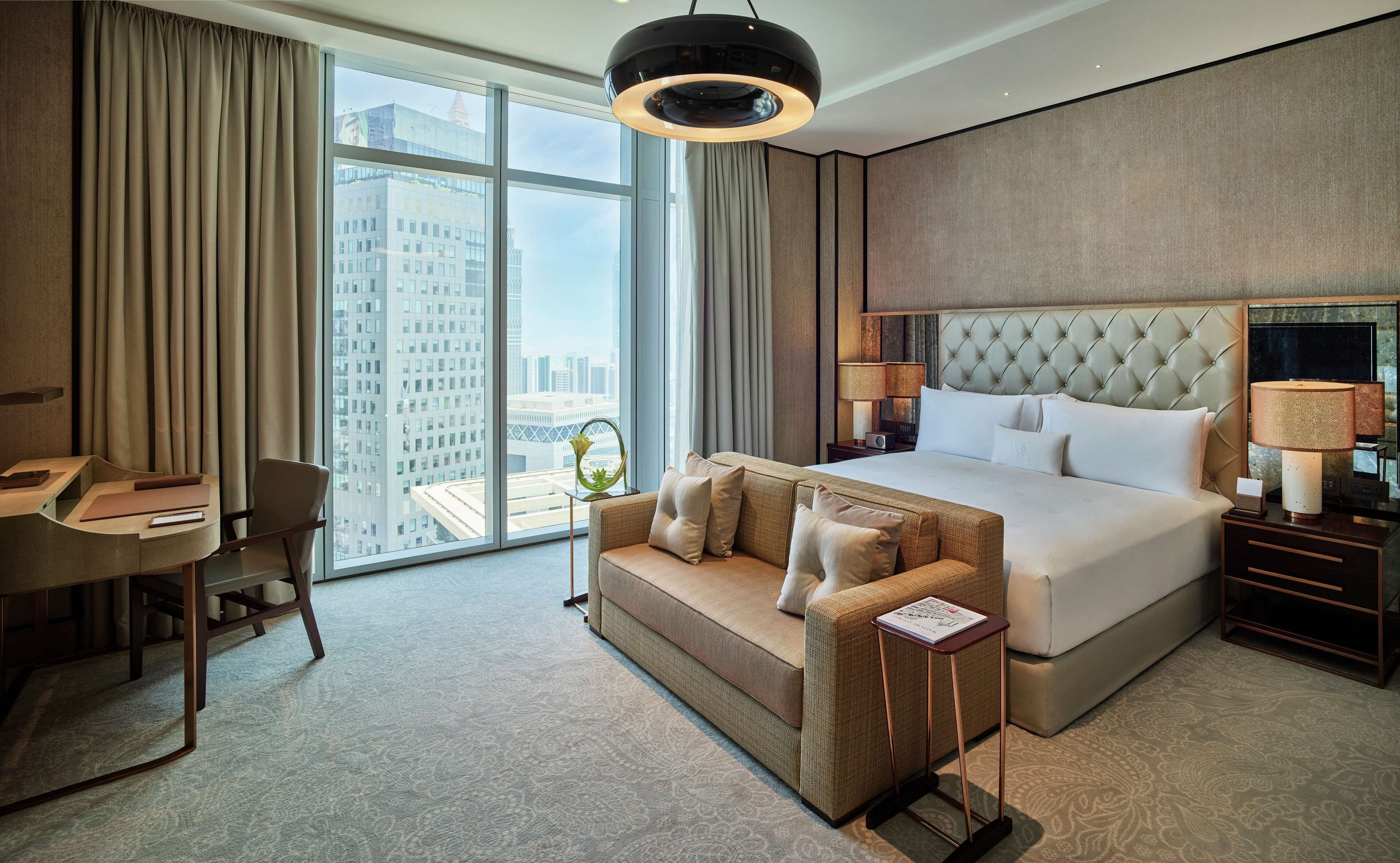 Una habitación de hotel moderna con una cama grande, un sofá, un escritorio y vistas de la ciudad a través de amplios ventanales.