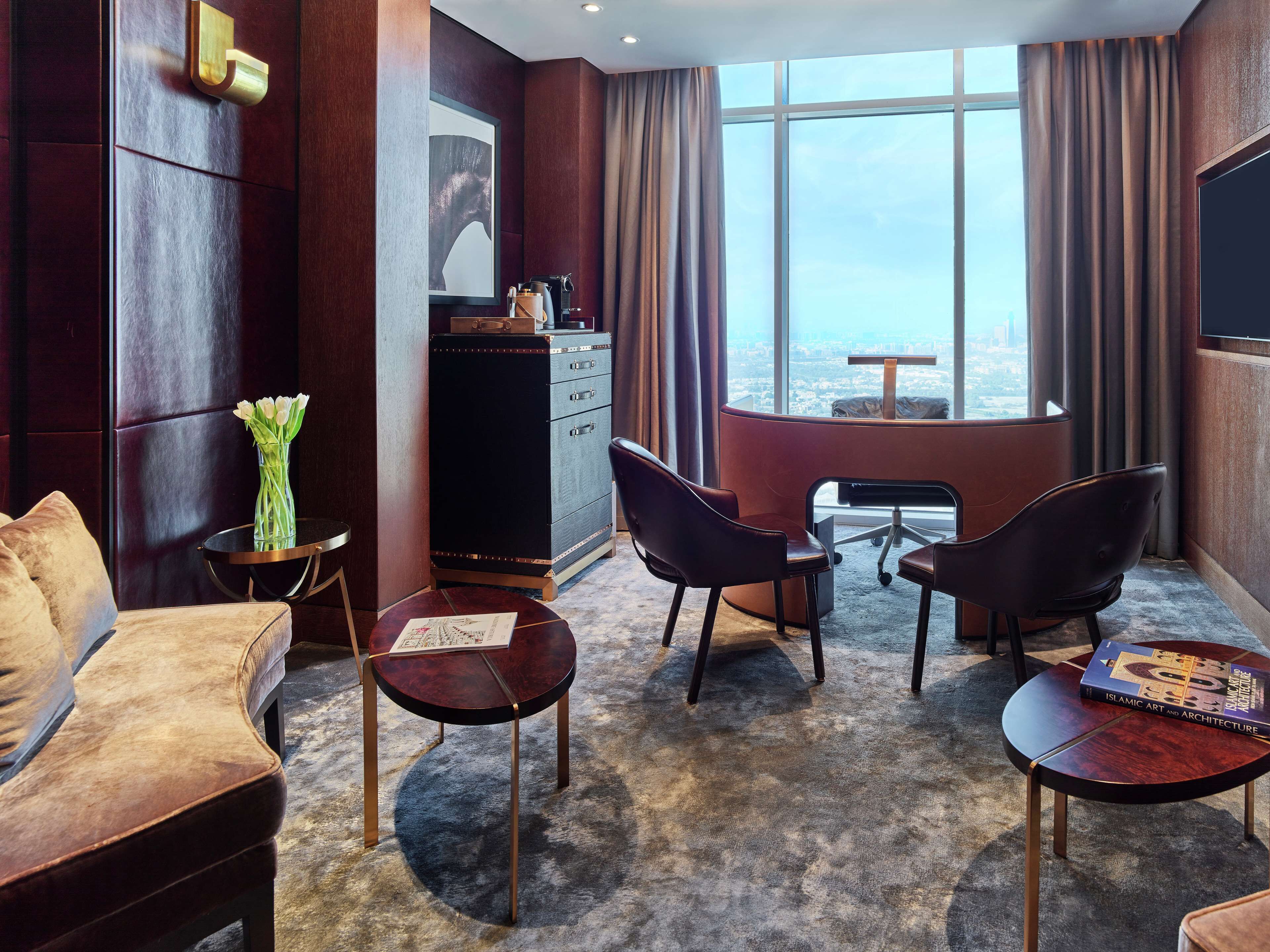 A modern hotel room with a large window, a sofa, two armchairs, a small table, and a cabinet.