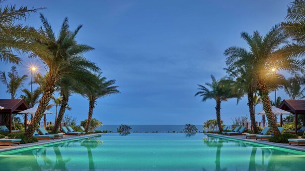 Zona de piscina con sillones de descanso y palmeras, cielo despejado y vista al mar.