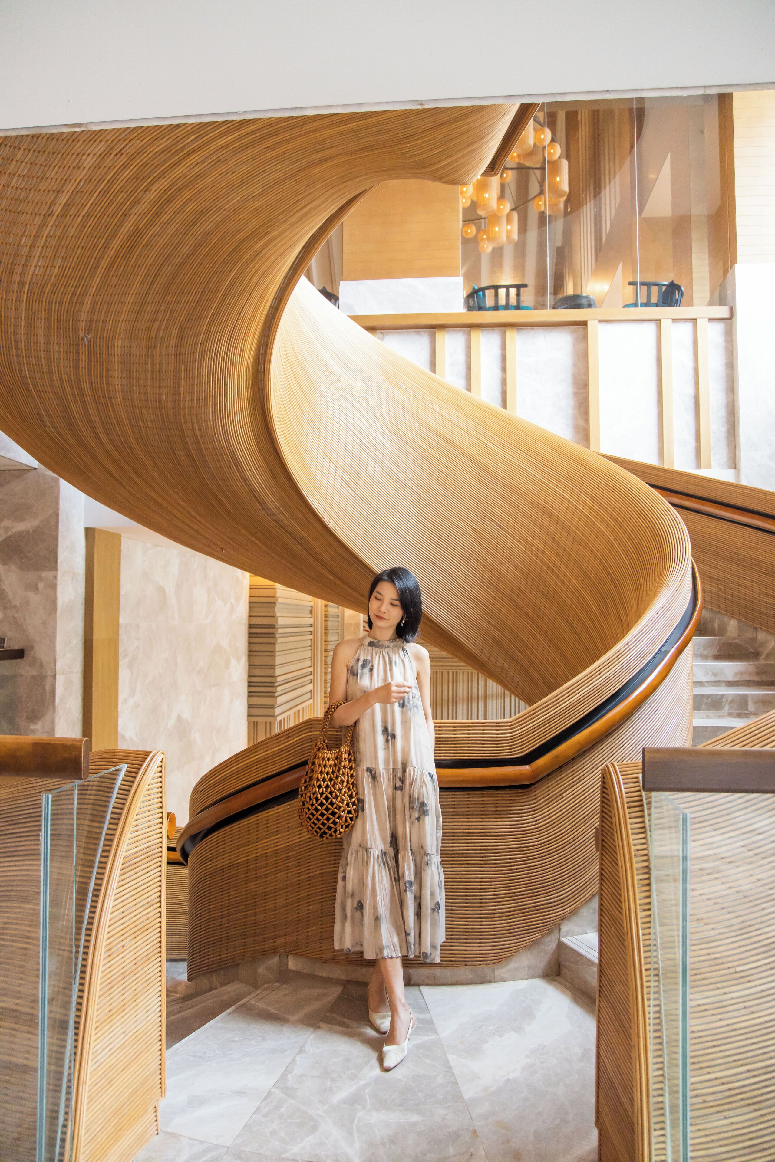 A woman standing on a curved wooden staircase with a modern interior design, including a glass railing and a hanging light fixture.
