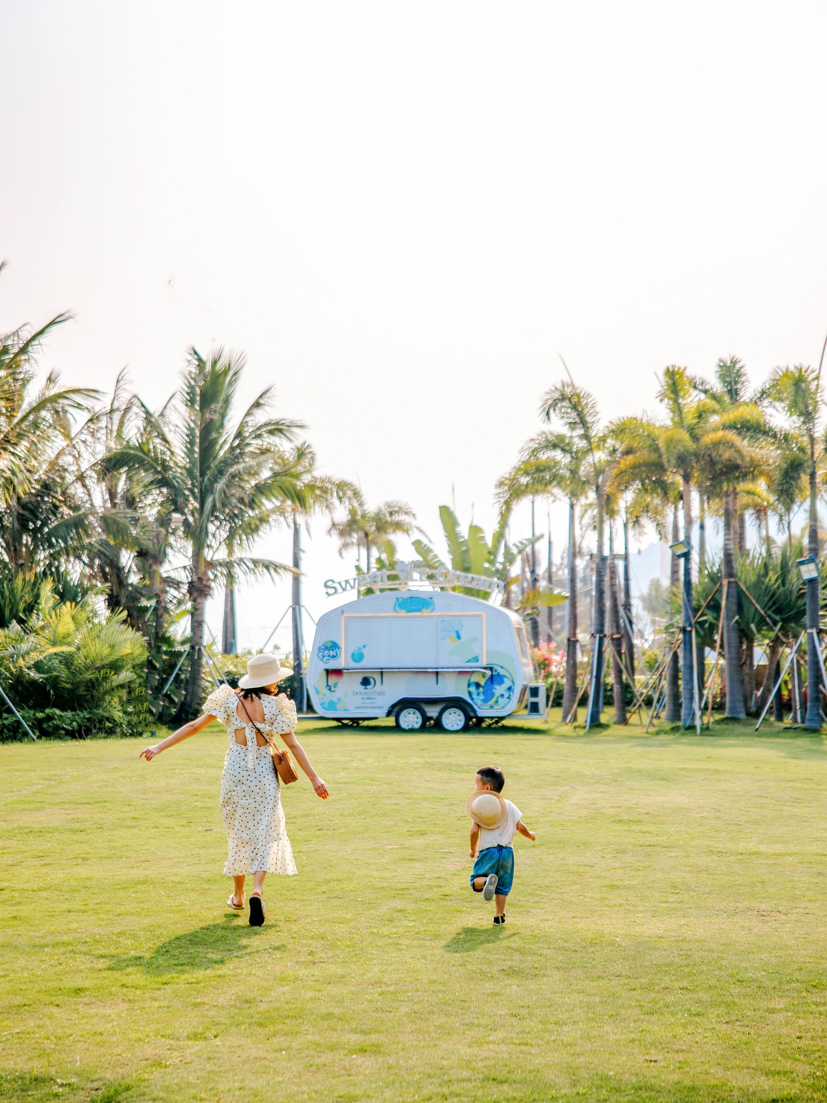A grassy field with a white trailer and a person in a polka dot dress and a child in a white shirt and blue shorts.