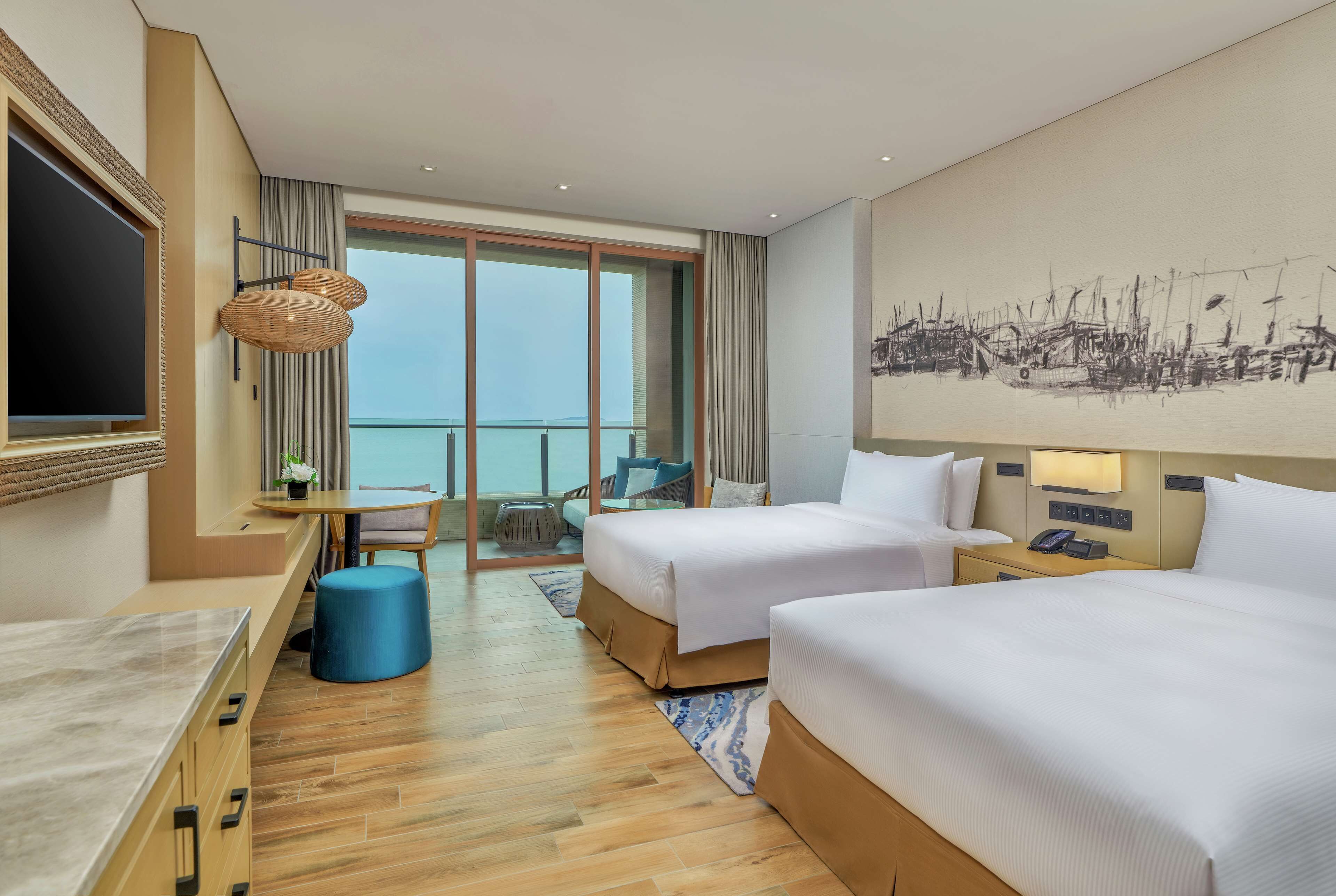 A hotel room with two beds, a large window, a dining area, and a view of the sea.