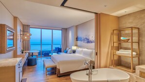 Una habitación de hotel moderna con una cama grande, un espacio para sentarse con vista al mar, una bañera y un estante de madera con toallas.