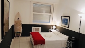 A hotel room with a bed, a red throw, a framed picture, a lamp, and a surfboard against the wall. - Jump in Bed Copacabana 3 - Estudio (Rio de Janeiro)