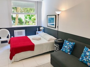 A hotel room with a bed, a sofa, a chair, a window with a view of greenery, a lamp, and a framed picture on the wall. - Jump in Bed Copacabana 3 - Estudio (Rio de Janeiro)
