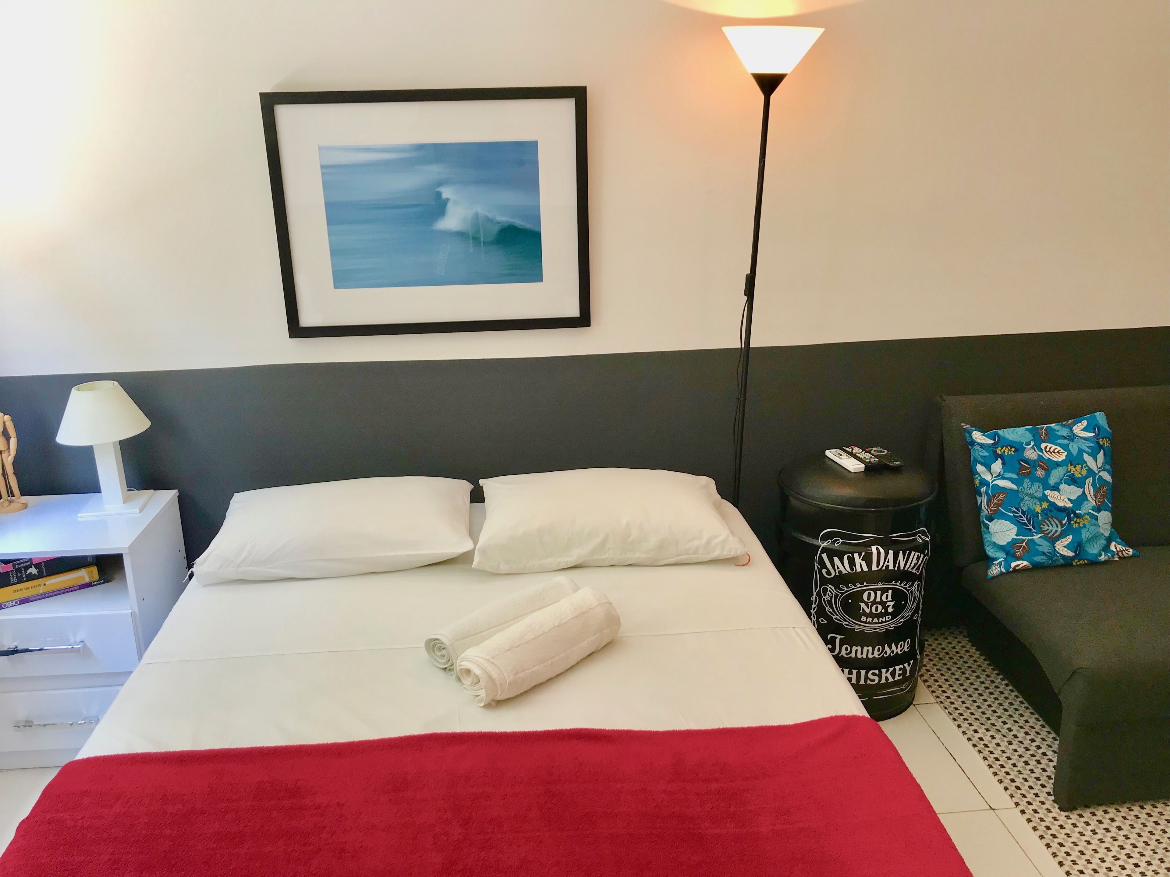 A hotel room with a bed, a nightstand, a lamp, a framed picture, a red blanket, a pillow, a small trash can, and a couch.