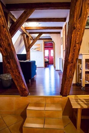 A loft-style room with exposed wooden beams, a staircase, a blue sofa, and a view into another room with a red wall.