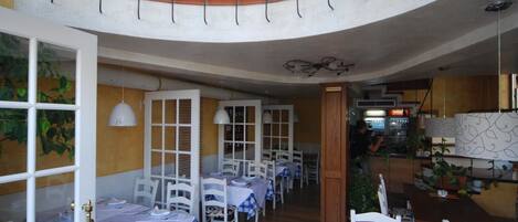 Un restaurante con mesas y sillas blancas, techo curvo y plantas en macetas.